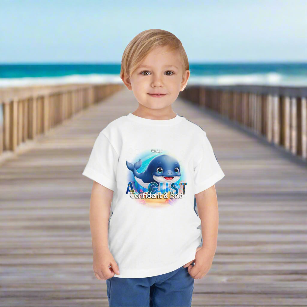 Toddler wearing kids birth month tshirt with August whale design standing on wooden boardwalk by the beach