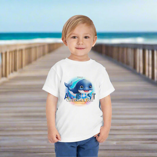 Toddler wearing kids birth month tshirt with August whale design standing on wooden boardwalk by the beach
